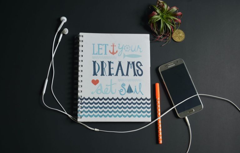 Notebook reading “Let your dreams set sail” beside a phone, earbuds, pen, and small plant