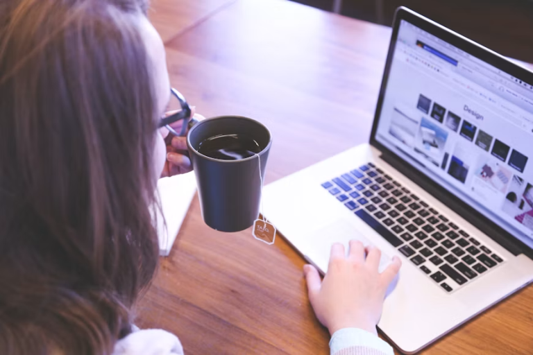 Person using a laptop with design images on screen while holding a mug