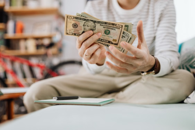 Person counting U.S. dollar bills beside a notebook and pen.