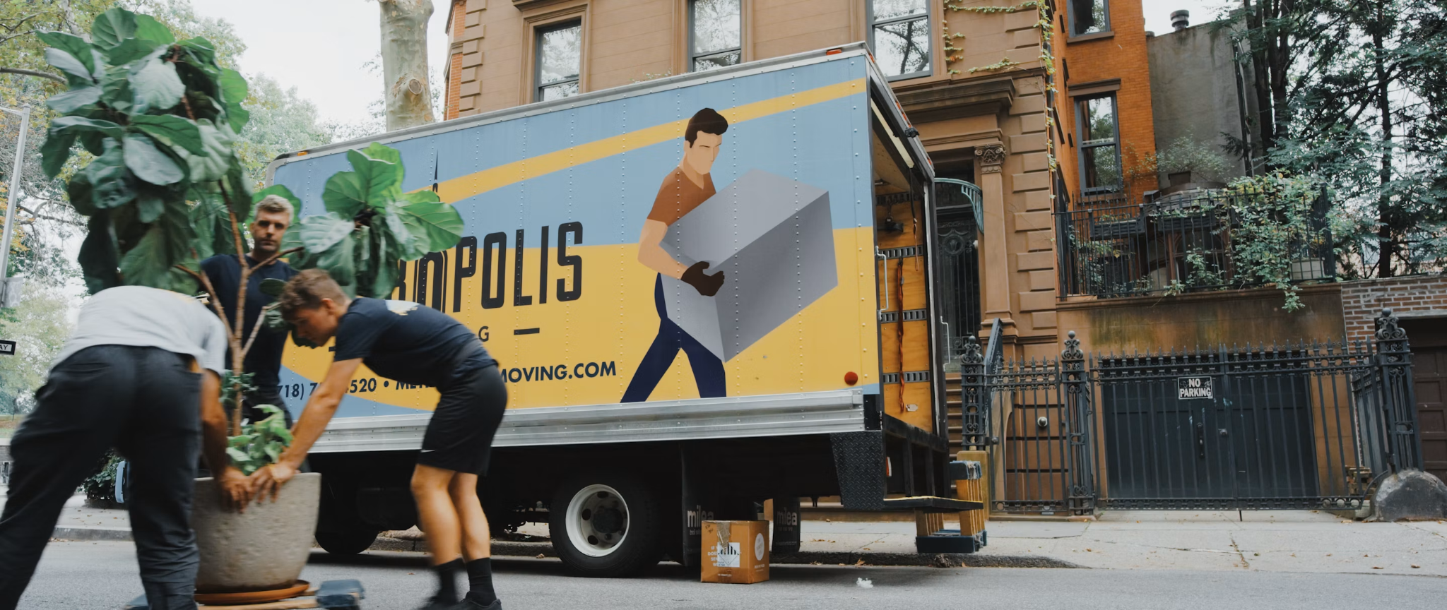 Movers loading a large potted plant beside a moving truck outside a residential building