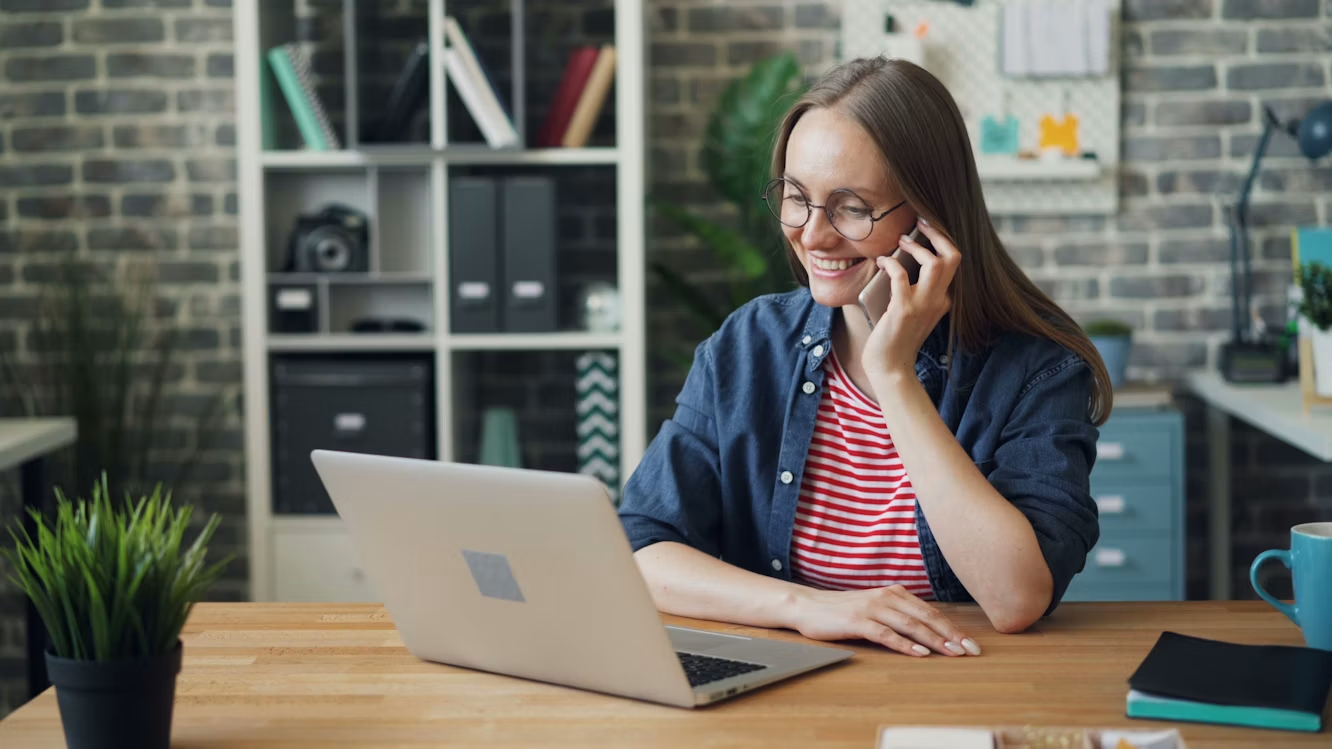 Woman speaking on a smartphone while working on a laptop in a home office