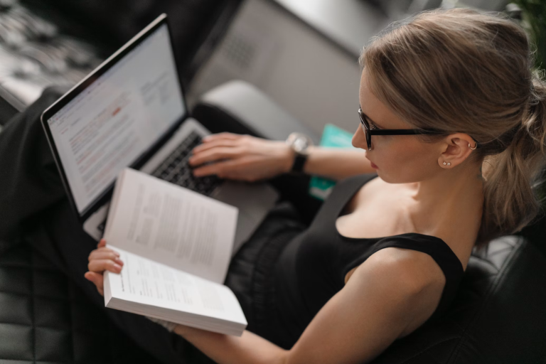 Woman reading a book while using a laptop for professional development