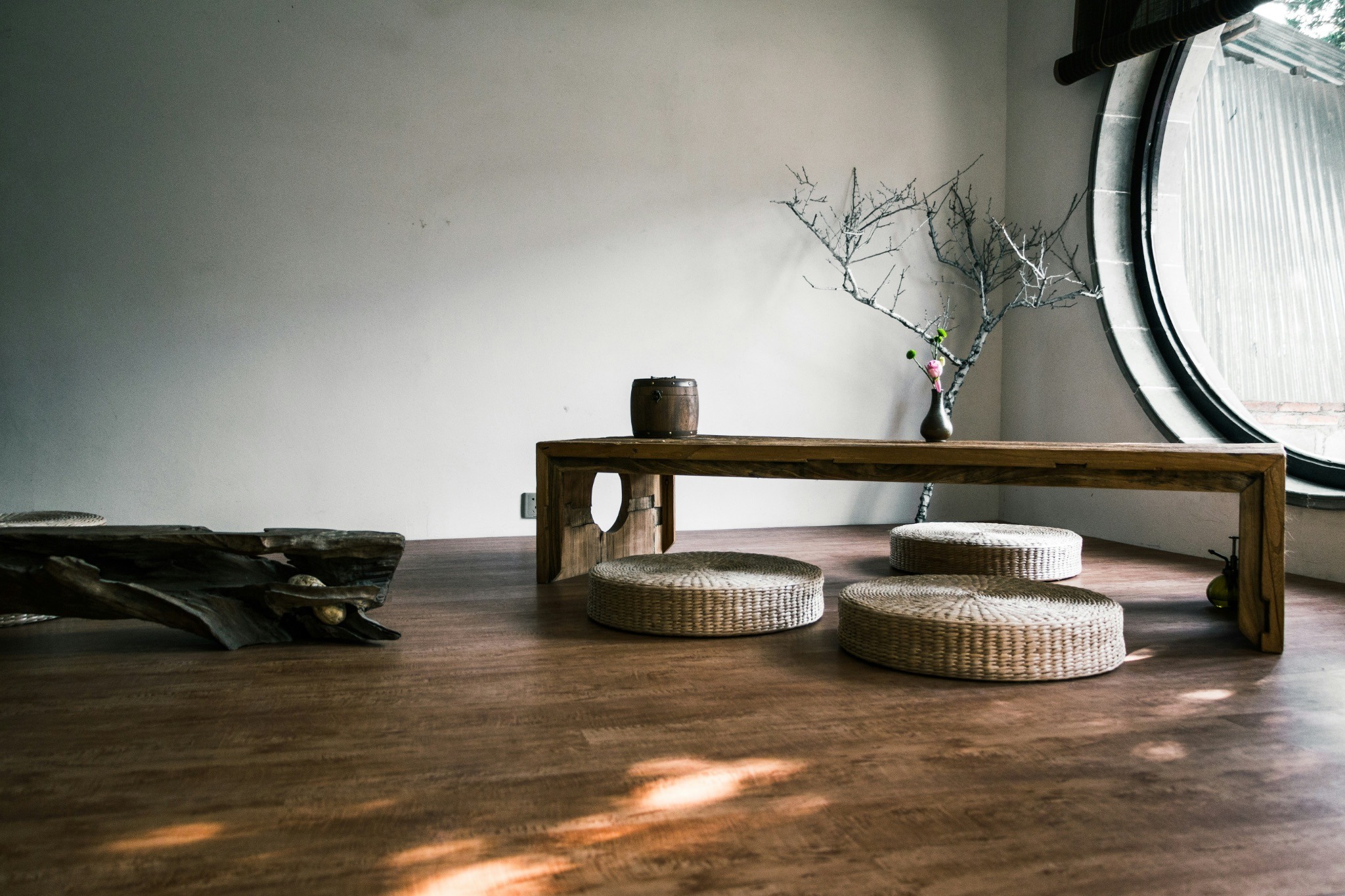 Minimal room with a wooden table, woven floor cushions, natural branches, and a large round window