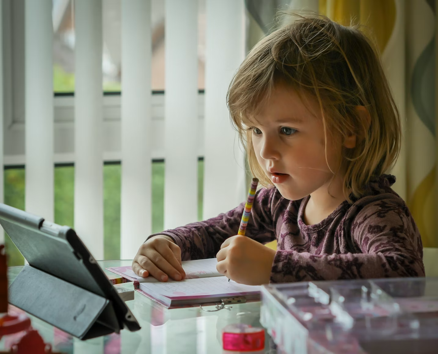 oung child writing in a notebook while watching a lesson on a tablet