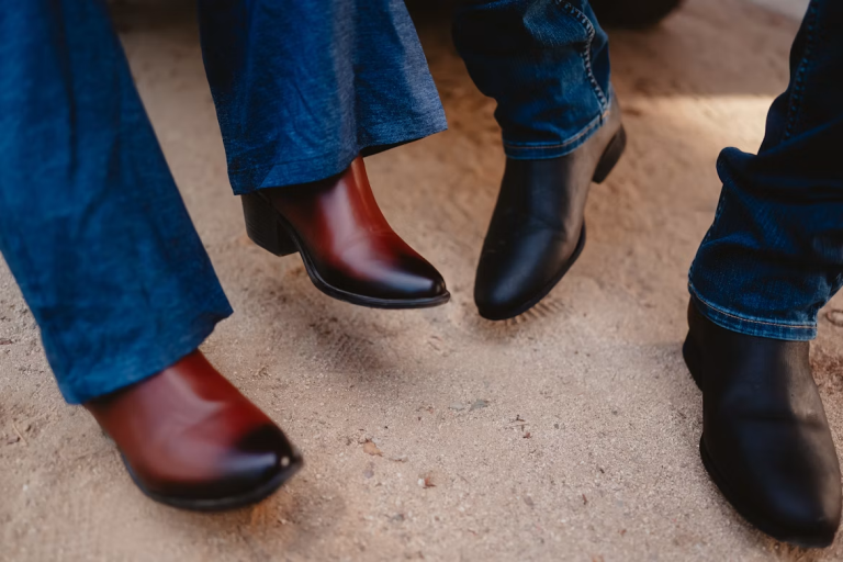 Three people wearing Western-style boots with blue jeans on sandy ground