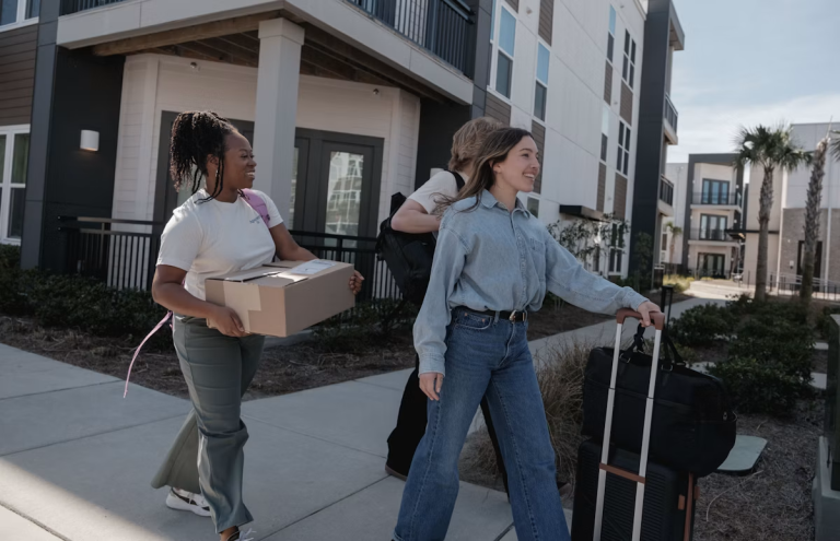Three women carrying a moving box and luggage outside a modern apartment building