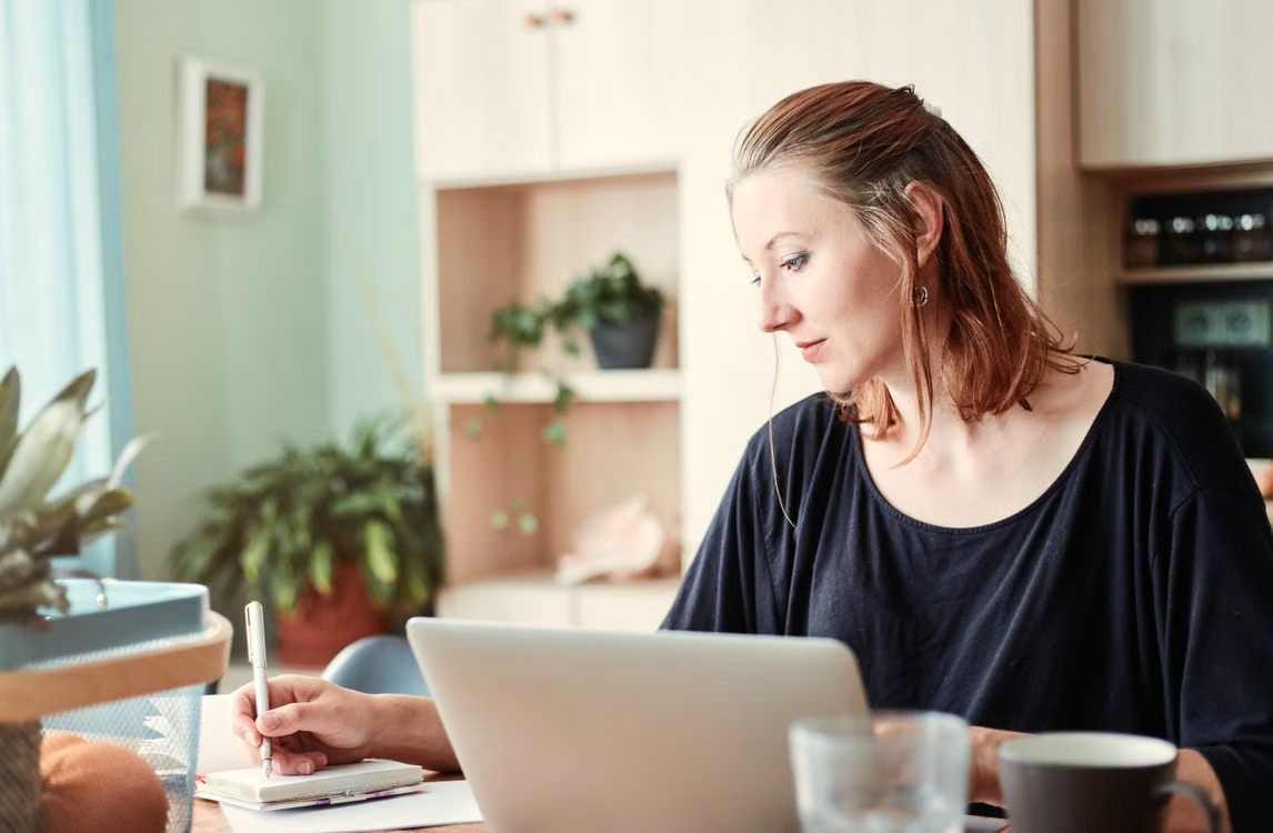 Woman taking notes while working on a laptop at home