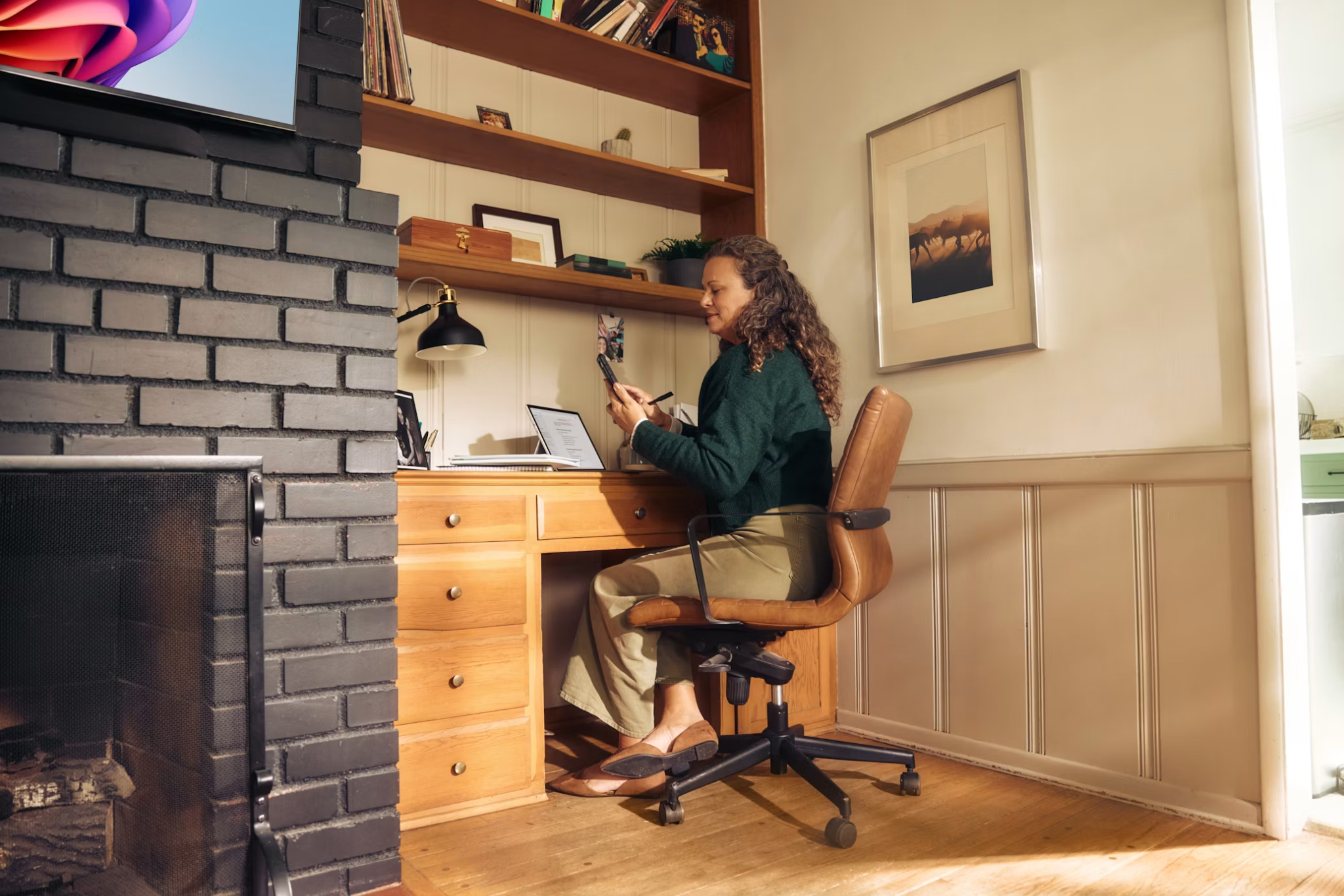 Woman using her phone and laptop at a desk in a sunlit home office