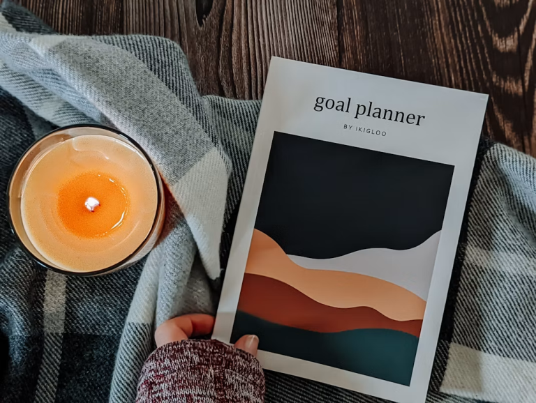 Hand holding a goal planner beside a lit candle and plaid blanket
