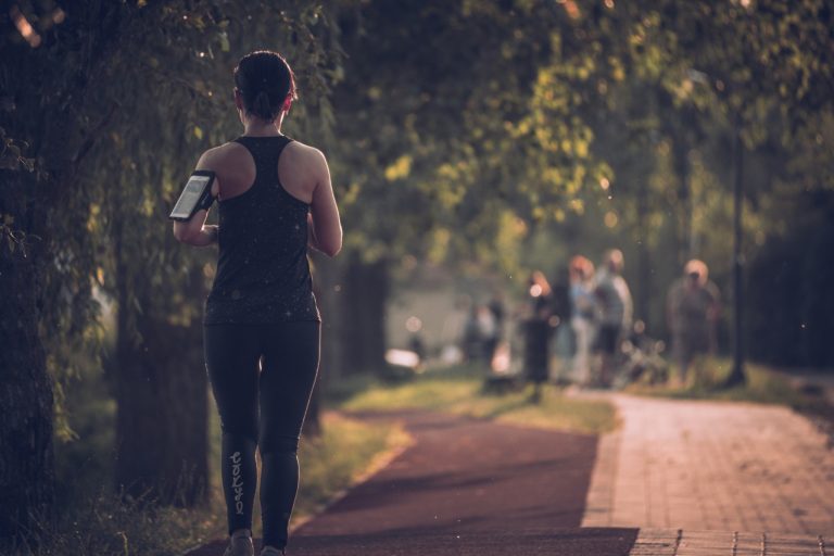 Woman walking on a tree-lined path while wearing athletic clothing and an armband phone holder.