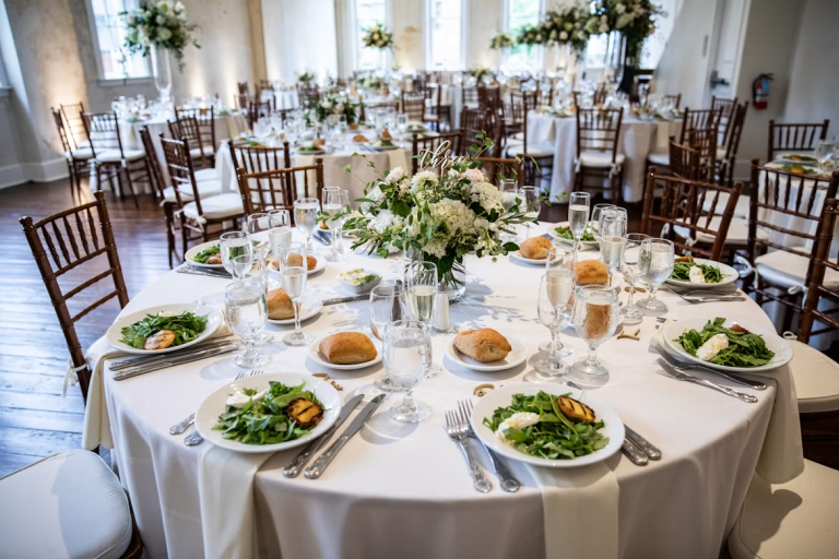 Elegant banquet tables set with white linens, floral centerpieces, place settings, and dinner plates in a bright event venue.