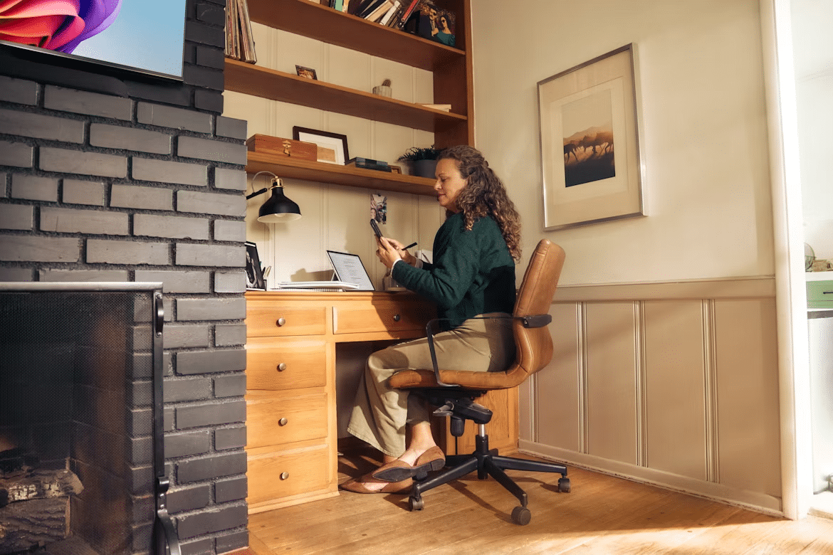 Woman working at a wooden desk in a cozy home office beside a fireplace.