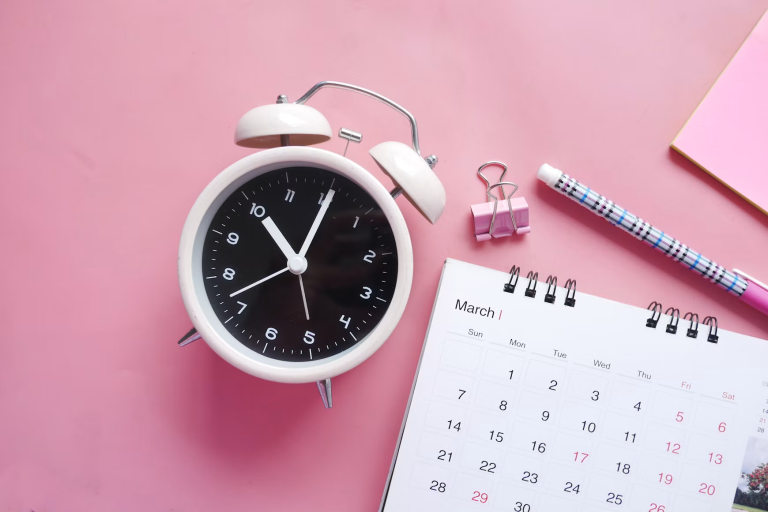 White alarm clock beside a monthly calendar and pencil on a pink background