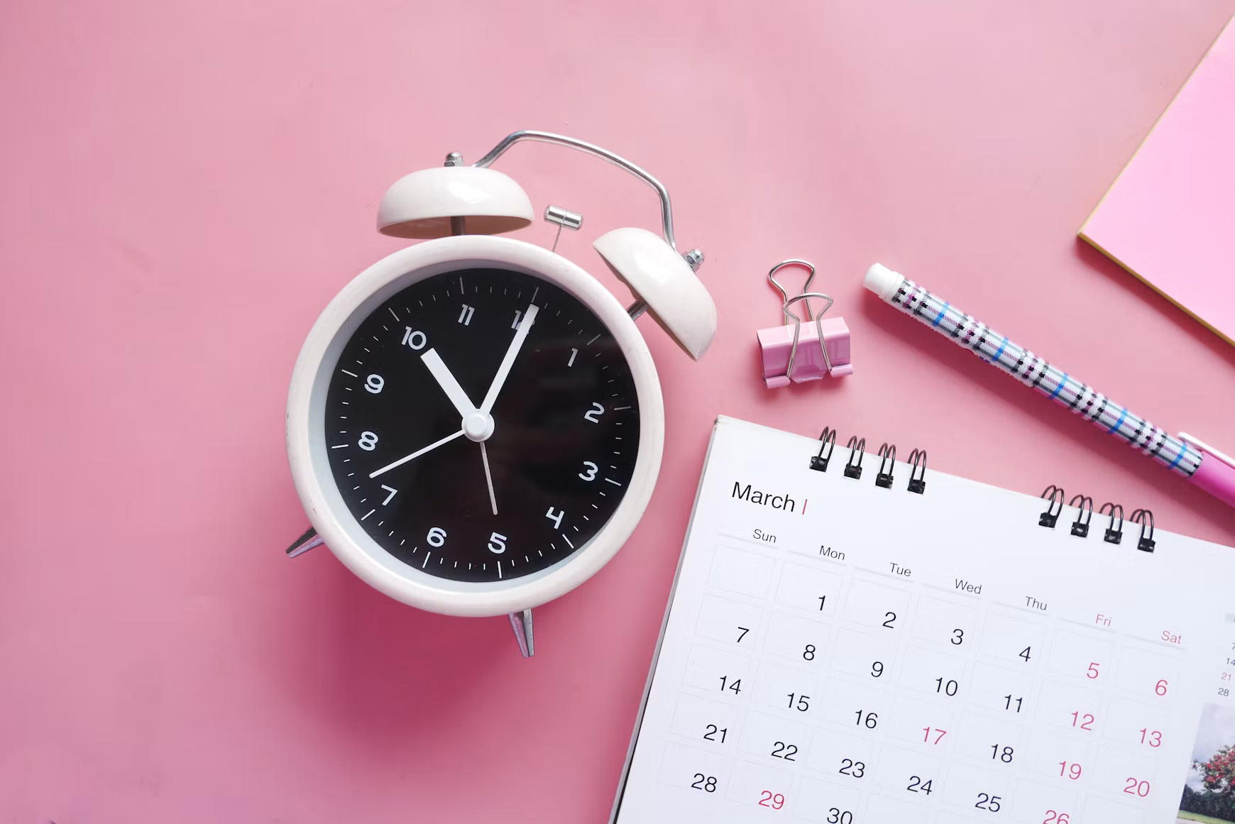 White alarm clock beside a monthly calendar and pencil on a pink background